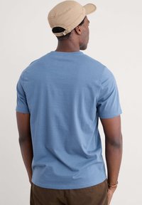 Ljust blå t-shirt med kort ärm i ett mjukt, slätt tyg. Har en klassisk rund halsringning och en avslappnad passform. Bärs tillsammans med bruna byxor.