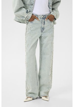 Osoba ubrana w jasnoniebieskie jeansy o szerokich nogawkach z perłowymi zdobieniami po bokach, biały top, dopasowaną kurtkę jeansową oraz białe buty z ostrym noskiem.