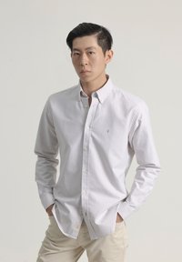 Camisa blanca de cuello con finas rayas verticales azules, diseño de botones, y un pequeño logo en el pecho, acompañada de pantalones beige claros.