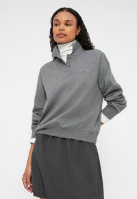 Γκρι πουλόβερ με ψηλό γιακά, δύο κουμπιά και ριχτές μανσέτες, συνδυασμένο με άσπρο μισό τσιγκέρι, παρέα με μια μαύρη φούστα.