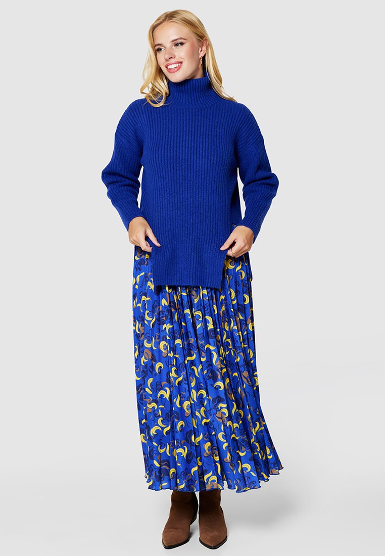 Niebieski sweter z golfem w prążki, zestawiony z lejącą się niebieską spódnicą z abstrakcyjnymi wzorami w kolorze żółtym i brązowym. Brązowe botki za kostkę.