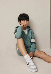 Hanorac cu fermoar verde mentă, cu dungi albe și bleumarin, asortat cu un set de pantaloni scurți; purtând adidași albi și șosete până la glezne.