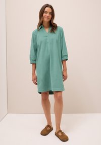 Abito di lino turchese con scollatura a V, maniche a tre quarti e aperture laterali, abbinato a scarpe slip-on marroni. Tessuto morbido e vestibilità rilassata.