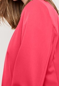 Mujer con cabello castaño claro hasta los hombros, vistiendo un blazer rosa brillante, vista de perfil contra un fondo liso.