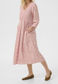 Nainen, joka wears pinkki kukkakuosisessa midimekossa, jossa on pitkät hihat ja taskut, yhdistettynä ruskeisiin tasapohjaisiin kenkiin, seisoo valkoista taustaa vasten.