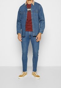 Uomo con giacca di jeans blu, t-shirt grafica color borgogna, jeans skinny blu e sneakers giallo senape, in piedi davanti a uno sfondo bianco.