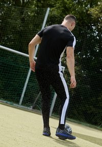 Muž v černém sportovním oblečení kopající do fotbalového míče na venkovním hřišti blízko branky se zeleným síťováním a stromy v pozadí.