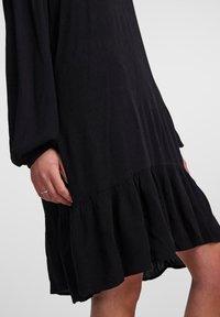 Vestido negro de tela texturizada, con mangas largas y un dobladillo de volantes en capas, con un corte relajado. Se puede ver un anillo de plata en la mano.