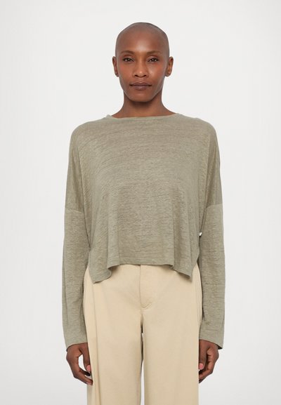 Top scurt cu mâneci lungi, de culoare verde măsliniu, dintr-un material texturat, cu un decolteu larg. Potrivit cu o pereche de pantaloni bej, lejeri.
