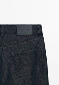 Jeans in denim scuro con una texture liscia. Presentano un'etichetta in pelle nella vita, una tasca posteriore e dettagli di cuciture sottili.