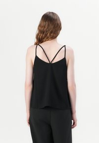 Femme aux cheveux bruns ondulés portant un haut noir sans manches avec de fines bretelles croisées dans le dos et un pantalon noir, debout sur un fond blanc.
