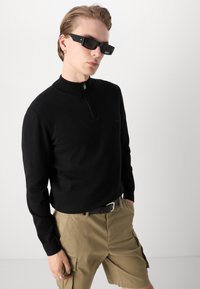 Pulover negru cu fermoar şi guler înalt, purtat cu pantaloni scurţi cargo kaki. Accesoriile includ ochelari de soare dreptunghiulari negri şi o curea neagră.