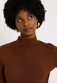 Pull en maille côtelée marron avec un col montant, présentant des lignes verticales et un motif en chevron sur l'avant. Texture douce et design ajusté.