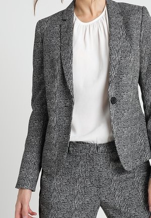 Kvinde iført en grå og sort ternet blazer og matchende bukser med en hvid bluse med rynket halsudskæring under.