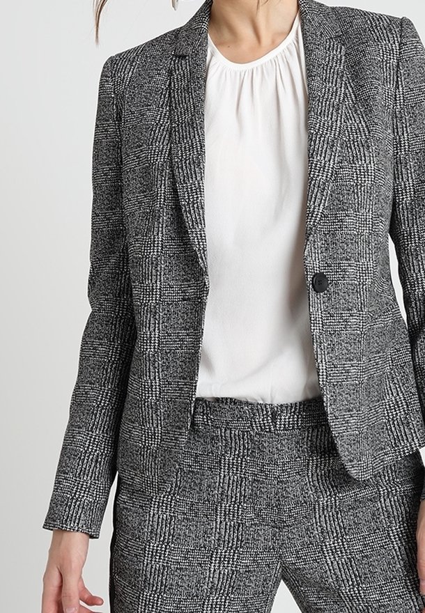 Femme portant un blazer à carreaux gris et noirs et un pantalon assorti, avec un chemisier blanc à encolure froncée en dessous.