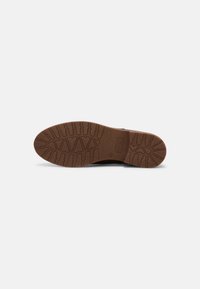 Semelle d'une chaussure marron avec des motifs de bande de roulement profonds et un talon légèrement surélevé, présentée sur un fond blanc.