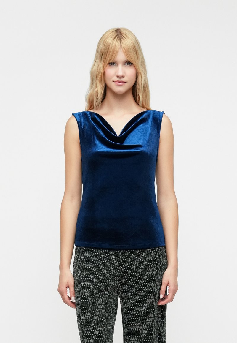 Navy blue, sleeveless top with a cowl neckline and a soft, shiny velvet texture. Worn with patterned black and white trousers.
