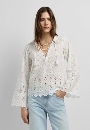 Vrouw draagt een witte blouse met vetersluiting, oogletdetails met bloemmotieven en geschulpte randen, gecombineerd met lichtblauwe spijkerbroek, tegen een effen achtergrond.