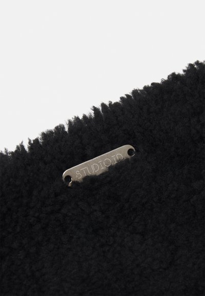 Tissu noir texturé avec une surface duveteuse, arborant une étiquette en métal embossée avec "STUDIOID". Le bord est soigneusement fini.