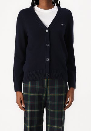 Personne portant un cardigan bleu marine boutonné sur une chemise blanche et un pantalon à carreaux verts, debout devant un fond blanc uni.