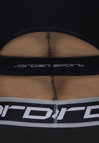 Svart idrettstopp med utskjæringsdetalj, med en tykk stropp merket "Jordan Sport" i hvit skrift. Glatt tekstur, tettsittende design.