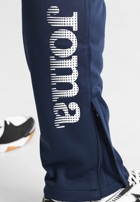 Marineblå sportbukser med hvid "Joma" logo trykt vertikalt. Har lynlås ved anklen og en tekstureret stofoverflade.