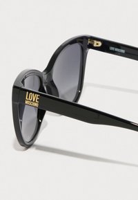 Črne sončne očala z sijajno površino, v obliki mačjih oči, s sivimi lečami in z zlatim logotipom "LOVE MOSCHINO" na levem ročaju.