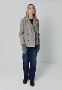 Abrigo gris de doble botonadura con solapas muesca, combinado con jeans azules y guantes de punto en color verde azulado. Botines negros completan el conjunto.