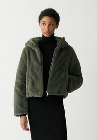 Olijfgroene faux fur cropped jas met een capuchon, ritssluiting en oversized mouwen; gedragen over een aansluitende zwarte ribbeljurk.