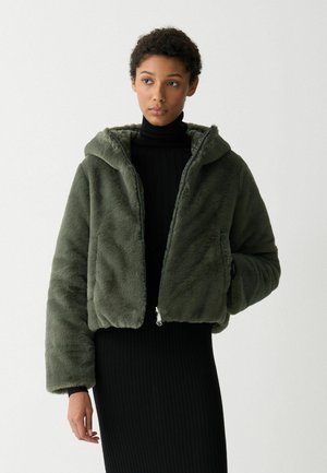 Olijfgroene faux fur cropped jas met een capuchon, ritssluiting en oversized mouwen; gedragen over een aansluitende zwarte ribbeljurk.