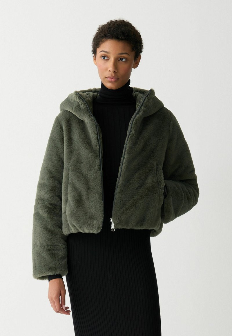 Olijfgroene faux fur cropped jas met een capuchon, ritssluiting en oversized mouwen; gedragen over een aansluitende zwarte ribbeljurk.