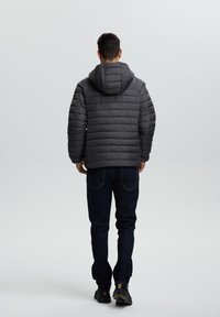 Mand der går væk iført en mørkegrå hættetrøje med fyld, mørkeblå jeans og sorte sneakers på en ensfarvet lys baggrund.
