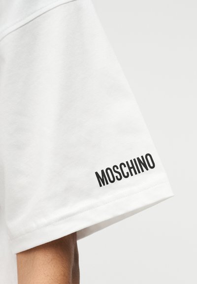 Λευκό βαμβακερό κοντομάνικο μπλουζάκι με μαύρο κεντημένο κείμενο "MOSCHINO" στο μανίκι. Απαλή υφή και κλασική κοπή.