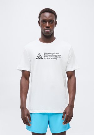 Homme portant un t-shirt blanc avec le texte et le logo « All Conditions Gear », associé à un short bleu vif, debout sur un fond blanc uni.