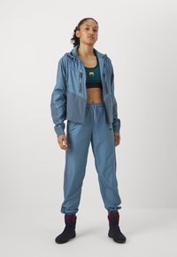 Casaco atlético azul-claro com calças de coordenado, costuras detalhadas, bolsos com fecho éclair e um top curto verde-escuro com um detalhe em dourado. Calçado preto.