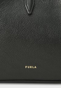 Melna ādas somiņa ar struktūru, kurai apakšā ir izteikts zeltains "Furla" logo. Minimālisma dizains ar elegantiem akcentiem.