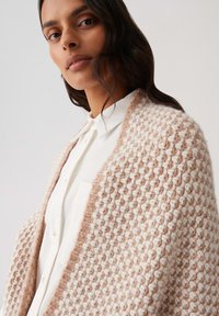 Strickschal in Beigetönen und Creme, mit einem strukturierten Muster. Über einem weißen Hemd mit Knöpfen getragen, wodurch ein geschichteter Look entsteht.