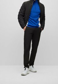 Persona con chaqueta negra acolchada, suéter azul con cremallera, pantalones negros y zapatillas blancas, de pie con las manos en los bolsillos.