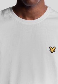 Witte t-shirt met korte mouwen en een ronde hals, met een klein geel en zwart vogel logo op de linkerborst. Zachte textuur.
