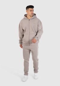Lys grå zip-up hættetrøje og matchende sweatpants lavet af blødt stof. Har hætte, lommer og hvide sneakers. Enkel, moderne design.
