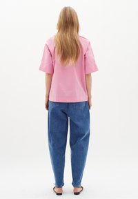 Kvinna med långt blont hår iförd en lös rosa kortärmad skjorta, blå högmidjade jeans och svarta sandaler med öppen tå, stående med ryggen mot kameran.