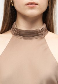 Νεαρή γυναίκα που φοράει αμάνικη μπεζ μπλούζα με ψηλό πιετέ γιακά και μακριά ίσια μαλλιά που φαίνονται στις πλευρές.