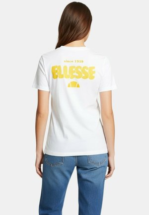 Femme aux longs cheveux bruns portant un t-shirt blanc avec un texte jaune "ELLESSE since 1959" et un jean bleu, vue de dos.