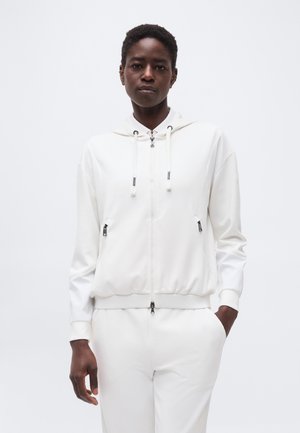 Femme portant un sweat à capuche blanc à fermeture éclair avec des cordons et un pantalon blanc, se tenant contre un fond clair uni.