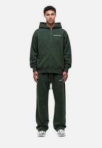 Felpa con zip verde scuro, dotata di tasche frontali e vita aderente, abbinata a pantaloni della tuta a vestibilità ampia. Dettaglio discreto del logo del brand.