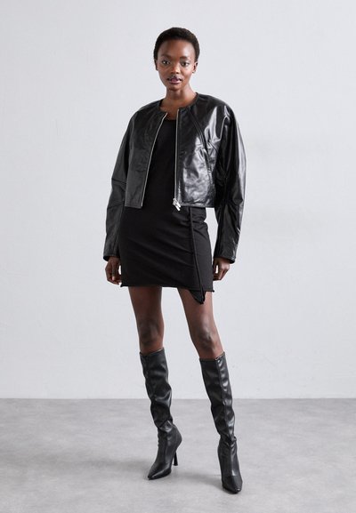 Fekete bőr bomber dzseki elülső cipzárral, fekete, testhezálló ruhával, amelynek oldalsó hasítéka van, és térd fölé érő, fekete magassarkú csizmával párosítva.