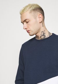Námornícky modrý sveter s okrúhlym výstrihom a dlhými rukávmi. Hladká textúra, s bielym akcentom na spodnej časti, minimalistický dizajn.