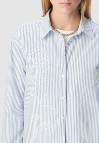 Camicia button-up a righe blu chiaro e bianche con ricami floreali bianchi sul lato sinistro. Presenta un colletto e una singola tasca sul petto.