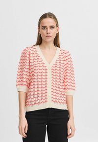Cardigan à manches courtes en crème avec un motif en vagues rose. Présente un col en V, une fermeture par boutons et un design en tricot texturé.