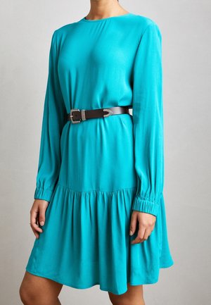 Kobieta ubrana w turkusową sukienkę z długim rękawem, z marszczonymi mankietami i brązowym paskiem z srebrną klamrą w talii.